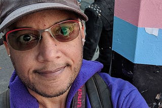 Selfie of Pax wearing a faded black AIDS walk cap, red-rimmed tinted glasses, purple hoodie, and black camera strap reading “funcrunch” in red letters. They are standing next to a pole decorated in white, pink, and blue stripes. A person with bright pink hair is visible in the background.