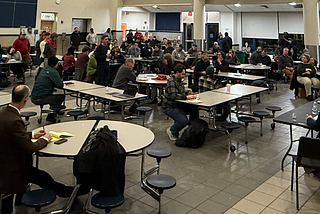 Nearly 100 people sit in a middle school cafeteria for a community meeting.