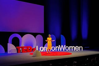 Anneka on stage wearing yellow dress, with arms opened out. Standing on big red circular carpet on a black stage. Behind are the letters ‘TEDxLondonWomen’ against purple-blue stage lights.