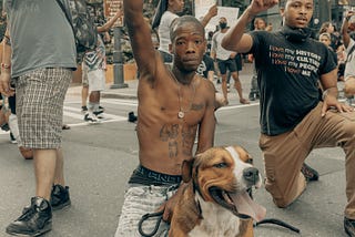 Thin shirtless Black Man with tattoos holds pitbull with one hand and makes a Black Power fist with the other hand.