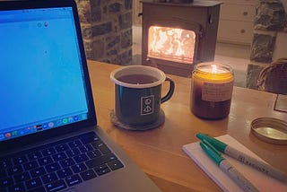 Laptop, mug of tea, candle, sharpies and notebook next to a log burner.