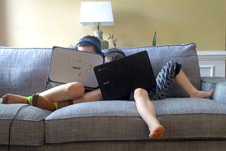 Two kids snuggling on a couch playing on their laptop computers.