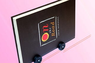 A copy of the book “On the Road” by Jack Kerouac is turned on its side and has racing wheels from a pinewood derby kit attached. The book is brown and the background is pink.