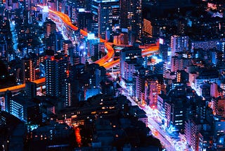 Night-time cityscape of high-rises as far as the eye can see. The buildings twinkle in iridescent blues and violets. Bright rivers of white and amber stream through long-exposed streets. Look at the collective power of society when we work together in harmony and mutual support.