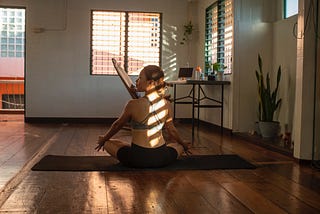 A woman sits cross-legged on a yoga mat in a tidy room with a desk and plants. The sun shines on her back in stripes through a window with blinds.