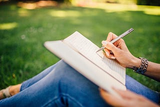 Woman’s hand writing in a notebook, sitting on a grass, wearing blue skinny jeans