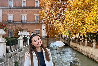 A woman wearing a long white coat stands on a bridge overlooking a canal with autum foilage in the background.
