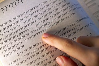 Image shows a finger pointing at a page in a book filled with question marks.
