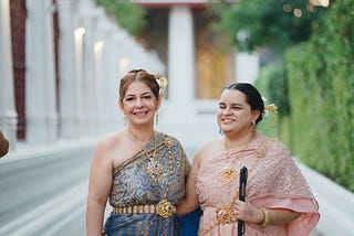 Dos mujeres están de pie y portan vestimenta tradicional tailandesa mientras comparten un abrazo mirando hacia la cámara. Una de ellas usa un bastón.