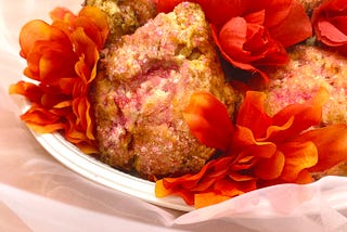 Scones with pink sanding sugar sitting on a white plate with orangey-red flowers dotted between the scones. Sitting on a soft peach background. Inspired by the Featherington sisters