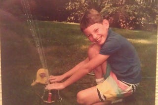 Young smiling fair-skinned boy in summer clothing with his hands in the jets of a portable lawn sprinkler