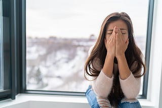 Anxiety, depression, worry concept — a photo of a woman sitting next to a window, covering her face in destress