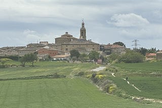 Verdant green fields lead to a town with a church spire rising over all the other buildings. There is a path running through the field and one person is walking towards the town.