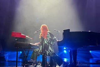 An image of singer-songwriter Tori Amos onstage in Seattle, wearing a green spangled outfit while playing her Bösendorfer piano with her left hand as her right plays a synthesizer.