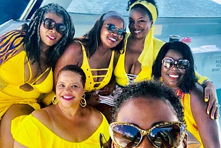 Six black women dressed in yellow swimwear and sunglasses on a boat, smiling.