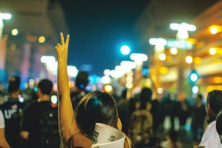 A young woman at the back of a crowd of people showing the peace sign.