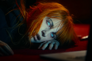 A woman with orange hair and bangs — Hayley Wiliams — lies on the floor in front of a laptop screen. Her eyes are glazed over, appearing white.