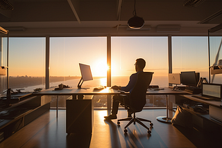 An executive working at a glass desk facing large glass windows near sunset hour.