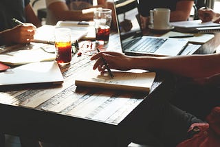 A group of people collaborating at a rustic wooden table with open laptops, notebooks, and drinks. Sunlight streams in, creating a warm, focused atmosphere ideal for a startup meeting or creative brainstorming session.