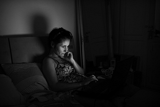 photo of a middle-aged woman sitting on her bed in the dark, staring at a laptop computer, her face illuminated by light from the computer screen