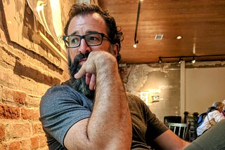 The author with dark curly hair, glasses, and a salt-and-pepper beard sits indoors against an exposed brick wall. He is wearing a grey t-shirt and resting his chin thoughtfully on his hand, looking off to the side. The warm-toned room has a wooden ceiling and worn plaster walls, with other people seated in the background.