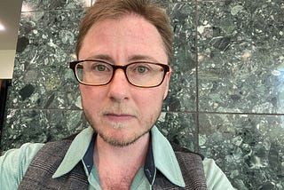 The author has short hair, a beard, and brown framed glasses. He’s wearing a green dress shirt, a grey vest, and is standing in front of a green marble wall. He has a serious face and is looking at the camera.