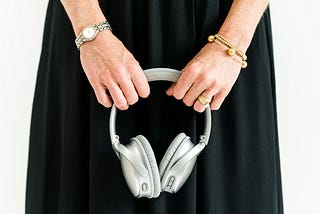 A close-up shot of a mature woman’s hands holding a silver headset. She wears a black dress, a silver watch on one wrist and gold bracelets on the other. She has gold wedding rings on her ring finger.