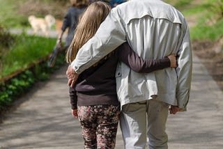A grandfather takes a walk with his granddaughter with their hands wrapped around each other.
