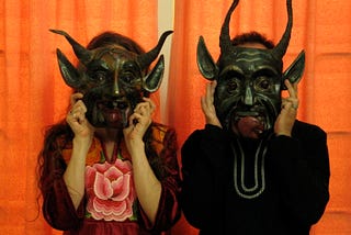 A man and woman hold Mexican devil masks in front of their faces.
