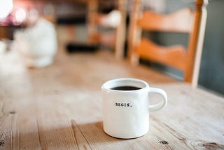 A white coffee mug sits isolated on the table. The coffee nearly reaches the top. One word is on the front in bold black letters. BEGIN.