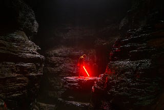 A man with a lightsaber, that's glowing red, in a dark cave. The mood is brooding.