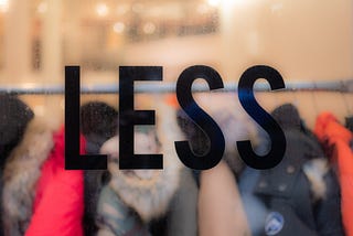 “The word “less” on a window, in focus with clothes behind it. Commentary on consumerism and waste in the fashion industry.”