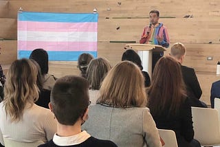 Dan Cantiller, wearing a multicolour rainbow hoodie, speaks to seated attendees from podium, beside a large Trans Pride Flag.