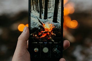 A hand holding a smartphone taking a photo of an outdoor campfire.