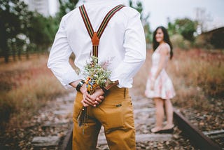 In the background of the picture, a woman wearing a sun dress is walking down a railway line. She’s smiling and looking back toward a man standing in the foreground. He is wearing a white shirt, brown trousers and braces. His hands are behind his back, and he is holding a bouquet of wild flowers.