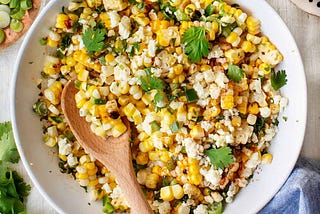 A bowl of Mexican street corn salad with cotija cheese, lime, green onion, and cilantro on a striped cloth.