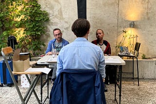 In the center foreground, a man sits facing away from camera and towards two other people. Those people are facing the man as they sit on opposite sides of a curtain, unable to see each other.