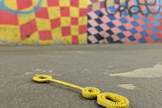 A discarded yellow bubble wand lies on the ground of a NYC subway tunnel. Brilliant painted checkerboard walls with graffiti in the background.