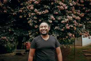 A man in a black shirt smiles in front of a tree.