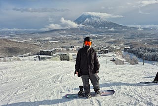 Author standing on a ski slope in snow gear with a snowboard strapped to one foot, you can see Niseko and Mt Yotei in the background