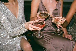 Women sitting in fancy cocktail dresses and holding coupe glasses of pink champagne.