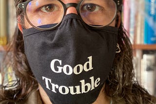 Close-up of me, wearing glasses, in front our bookshelf, my face half covered by a mask reading, “Good Trouble”.