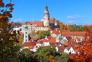 A circular tower, pale pink marble with faded colorful murals around it and a patinaed green bell tower. It stands among a jumble of mostly white buildings with brick-red rooftops. Trees are in full fall colors.