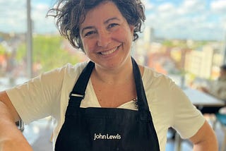 A picture of a smiley Chiara, wearing a white t-shirt and a black apron with the John Lewis brand on it.