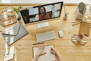 Shot taken from above looking down at a video call on a Mac screen sitting on a wooden surface. Stationary, keyboard, mouse, a book and hands surround the scene.