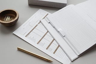 An agenda laid open on a desk, a gold pen, and a wooden bowl filled with gold paperclips.