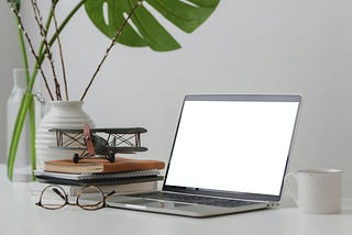 A macbook pro, a cup of coffee, books, glasses, and some plants on top of a white desk.