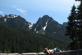 A woman lays on the wall of an overlook in Mt Ranier State Park, mountains, trees, and sky in the background