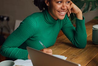 A young woman wearing a green turtleneck leans on a wooden desk with a notepad and laptop. Her face rests against her hand and she has a warm smile.