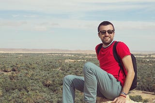 Man sitting in red shirt and jeans and sunglasses on a rock overlooking trees in background